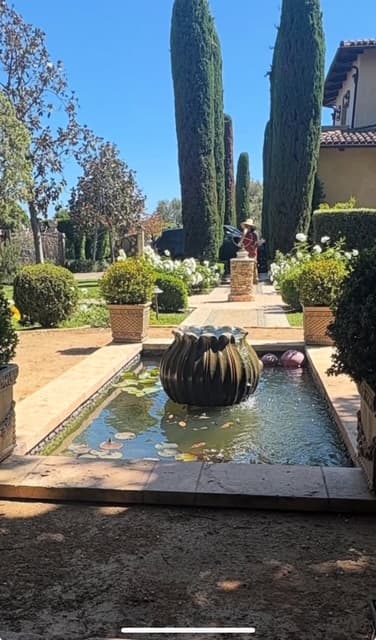 Rectangular garden pond with a ribbed fountain, surrounded by tall cypress trees and lush greenery.